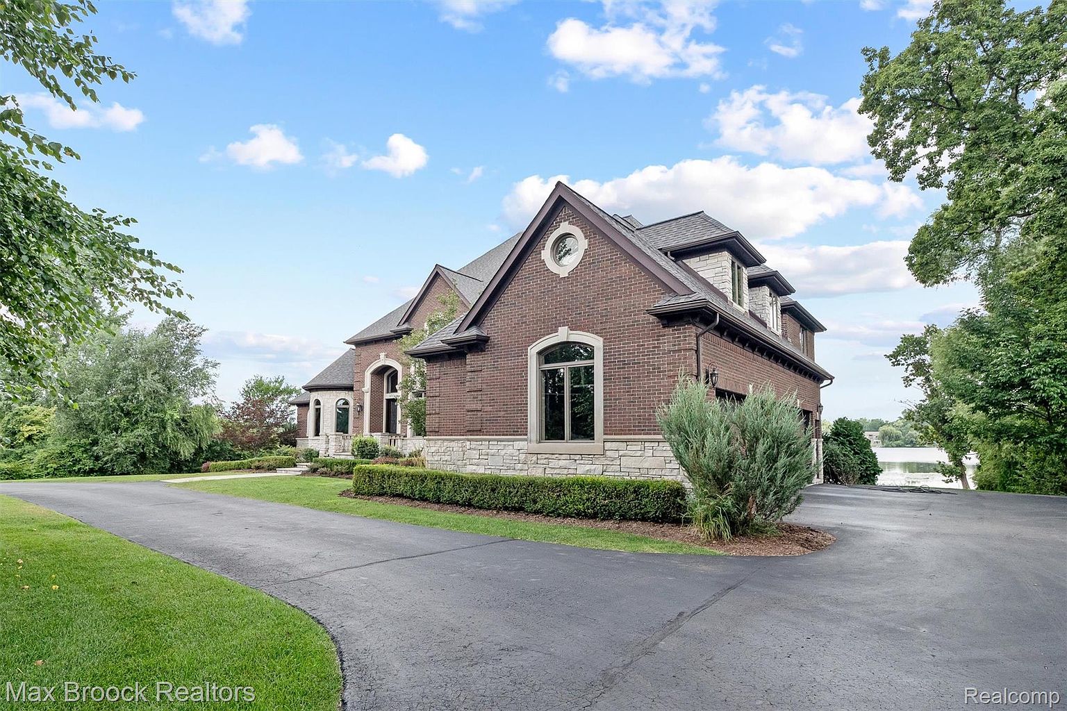 This is a side view of a large, luxurious brick home with stone accents and a well-manicured lawn. The house features multiple gables, arched windows, and a circular window detail. A paved driveway leads to the house, and mature trees and landscaping surround the property, suggesting privacy and a well-established neighborhood.