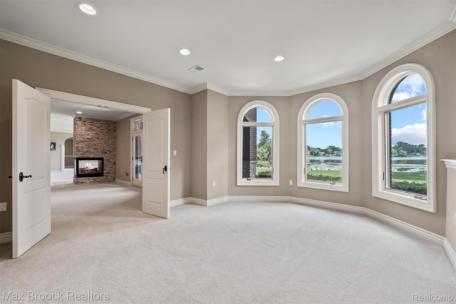 This is a spacious primary bedroom featuring three arched windows that offer a view of a lake. The room has neutral-toned walls, white trim, and plush carpeting. An open doorway leads to another room with a stone fireplace, adding a cozy and luxurious touch.