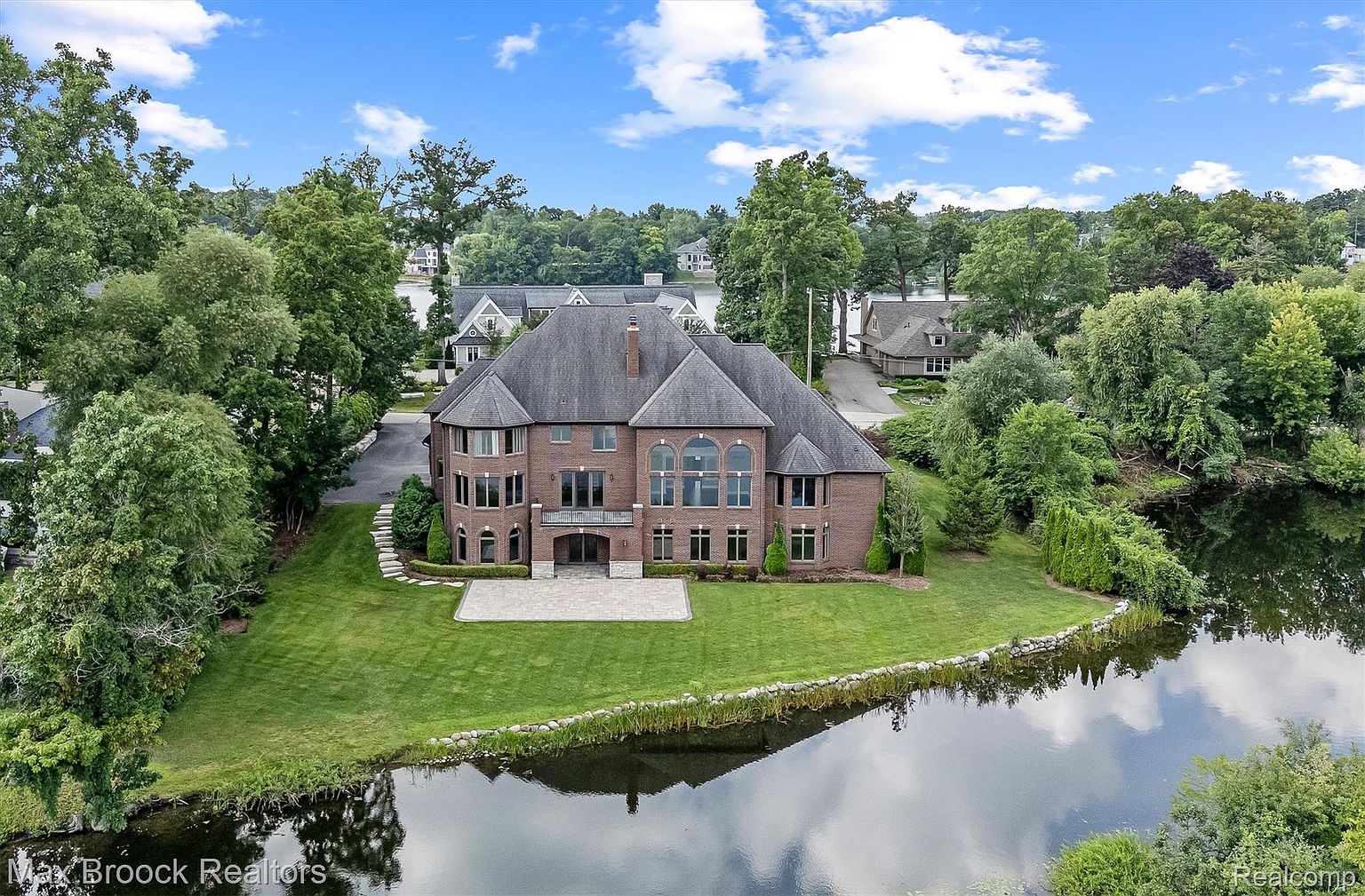 This aerial view showcases a large, luxurious brick home situated on a well-manicured lawn with waterfront access. The house features a complex roofline, multiple windows, and a patio area. The surrounding landscape includes mature trees and a serene body of water, enhancing the property's appeal and privacy.
