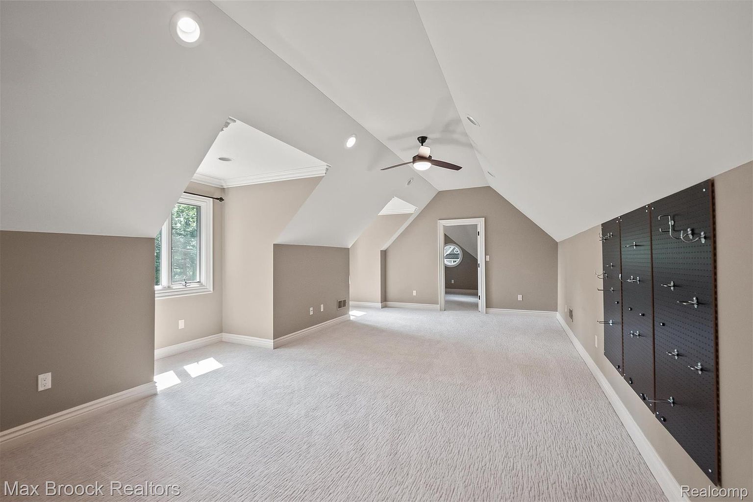 This is an interior shot of a spacious attic room, featuring a light gray carpet and neutral-toned walls. The room has a unique architectural design with sloped ceilings and dormer windows, creating a cozy yet open atmosphere. A ceiling fan and recessed lighting provide ample illumination, while a dark-colored pegboard wall adds a touch of modern functionality.