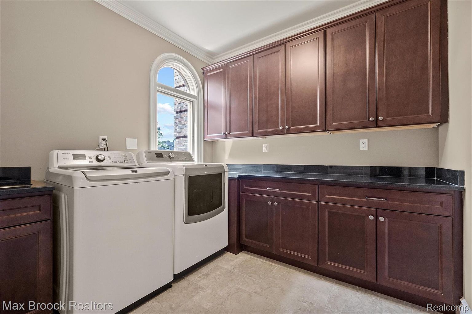 This is a well-organized laundry room featuring a front-loading washer and dryer set in white, complemented by dark wood cabinetry above and below a dark countertop. A large arched window provides natural light, and the neutral wall color creates a clean and functional space. The perspective is from the front, showcasing the appliances and cabinetry.