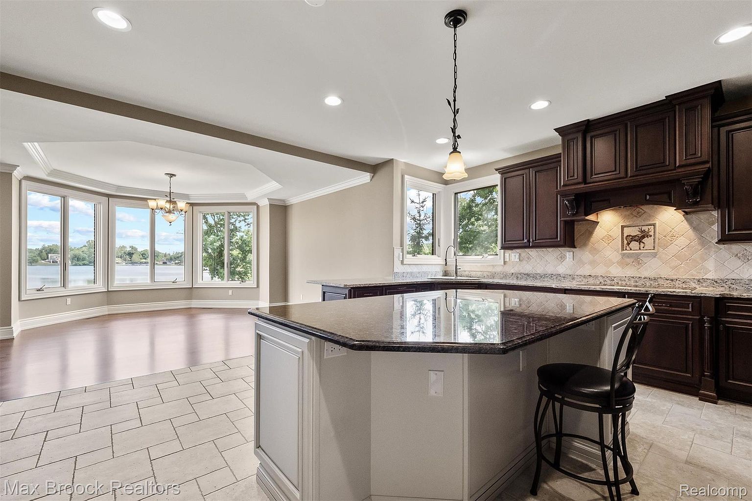 This is a well-lit kitchen featuring dark wood cabinetry, granite countertops, and a central island with seating. The kitchen opens to a dining area with large windows offering a view of the water. The flooring is tiled, and pendant lighting hangs above the island, creating a warm and inviting atmosphere.