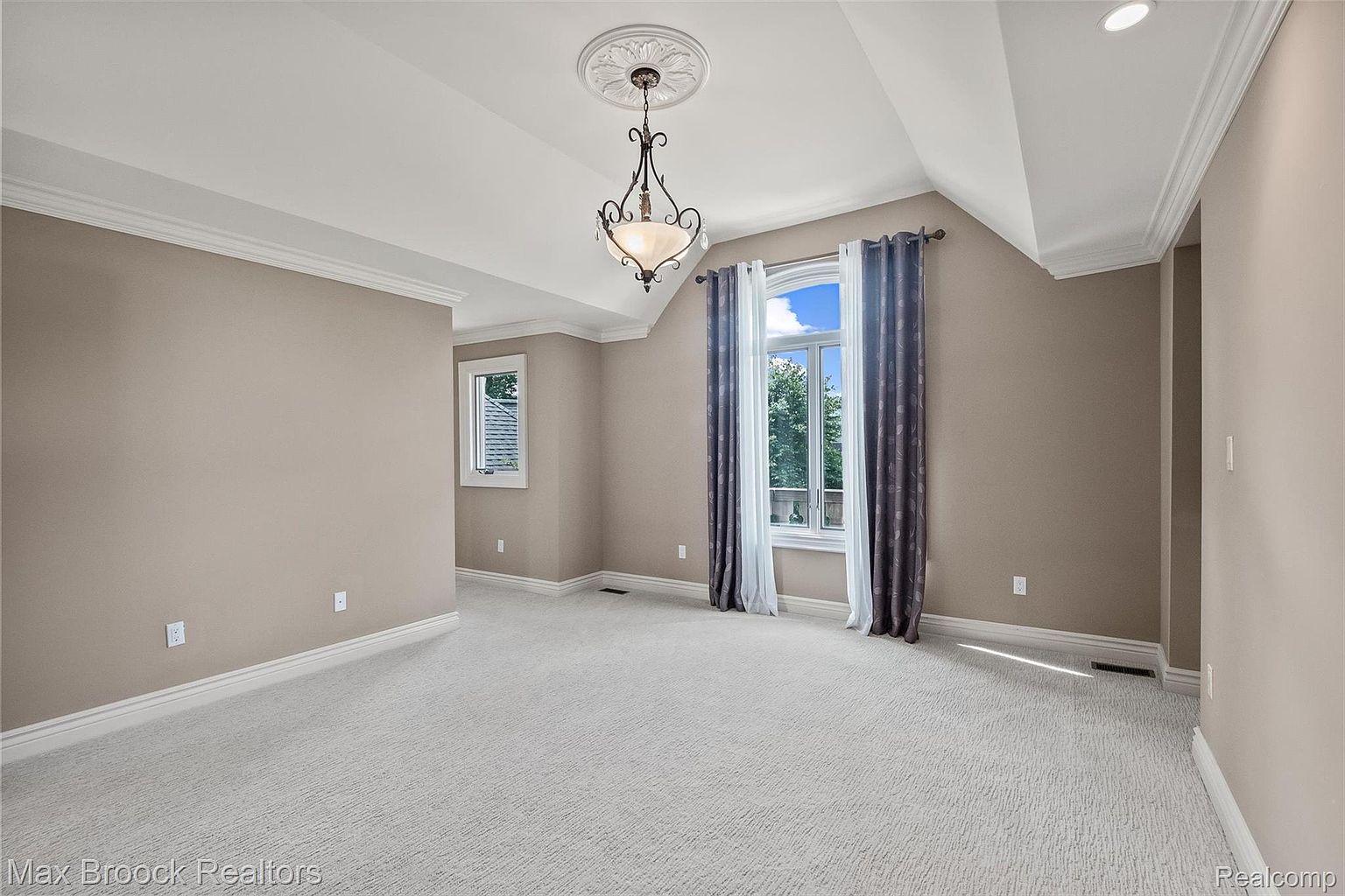 This is a spacious primary bedroom featuring neutral-toned walls and light-colored carpeting. A decorative chandelier hangs from the ceiling, and natural light streams in through a window adorned with elegant curtains. The room exudes a sense of calm and sophistication, making it an inviting retreat.
