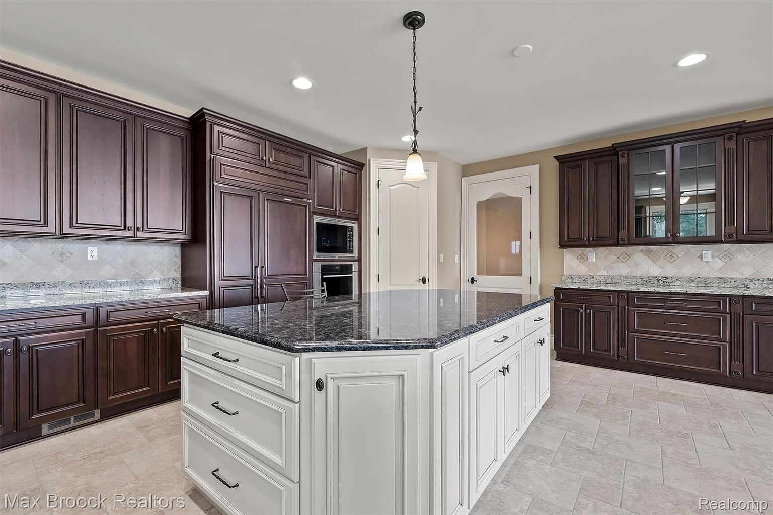 This is a well-appointed kitchen featuring dark wood cabinetry along the walls and a contrasting white island with a dark countertop. The kitchen includes built-in appliances and a tile backsplash. The flooring is a light-colored tile laid in a herringbone pattern, and a pendant light hangs above the island, adding a touch of elegance.