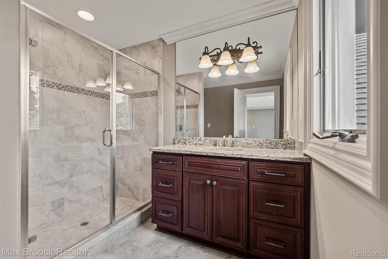 This is a well-appointed primary bathroom featuring a glass-enclosed shower with marble-like tile and a dark wood vanity with a granite countertop. A large mirror reflects a decorative light fixture, and the overall design exudes a sense of luxury and cleanliness. The perspective is from the doorway, showcasing the main features of the bathroom.