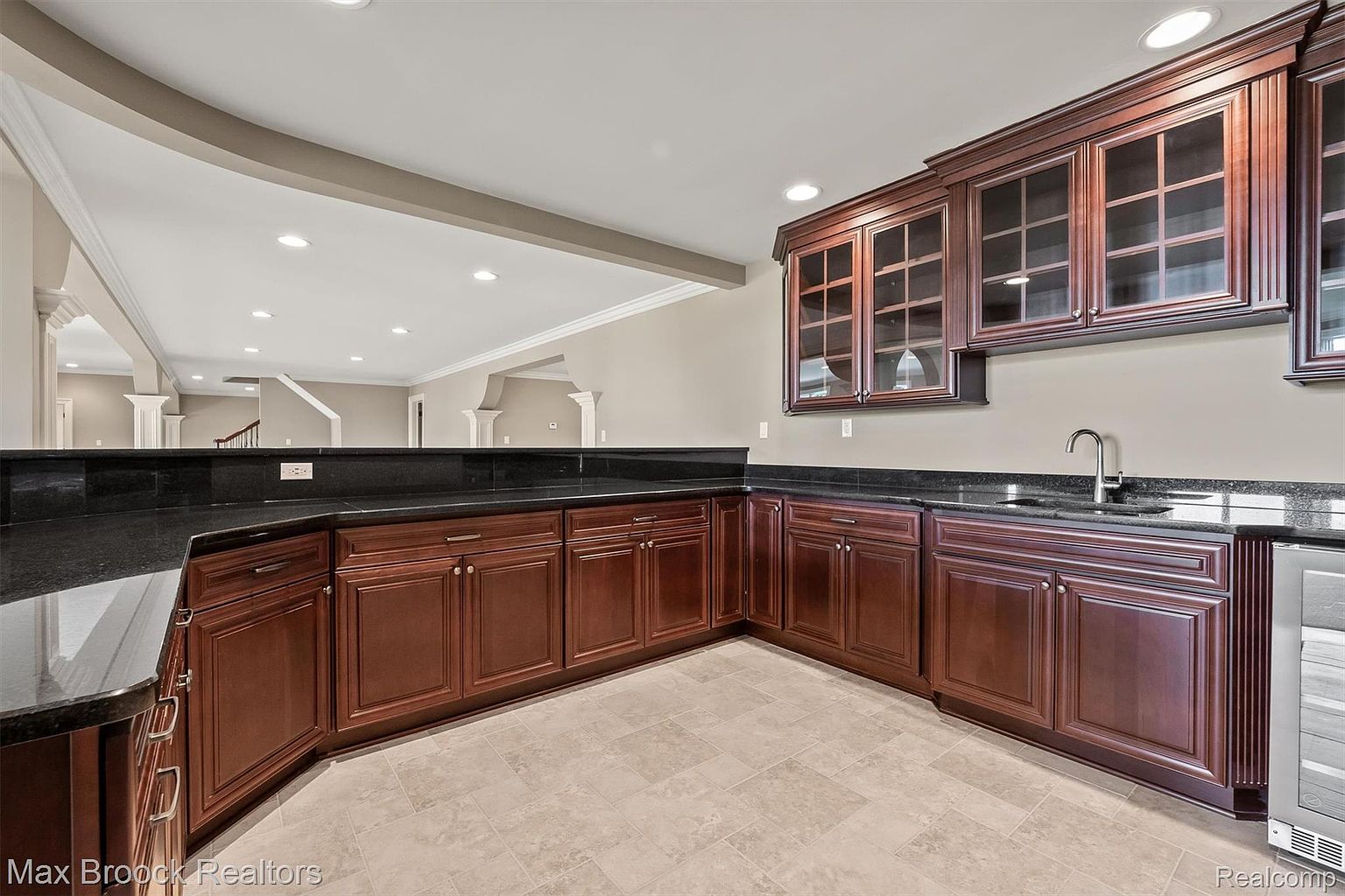 This is an interior shot of a basement wet bar area. The bar features dark wood cabinetry with glass-fronted upper cabinets, black granite countertops, and a stainless steel sink. The flooring is light-colored tile, and the space opens up to a larger basement area with columns and recessed lighting.