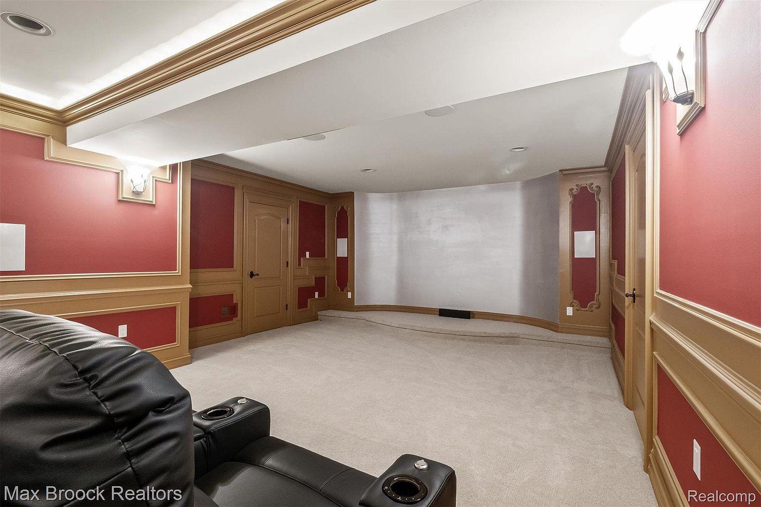 This is a finished basement space designed as a home theater or entertainment area. The walls are a combination of red and wood paneling, with a neutral carpet covering the floor. A large, comfortable-looking black leather seating arrangement is visible in the foreground, suggesting a relaxing and immersive viewing experience.