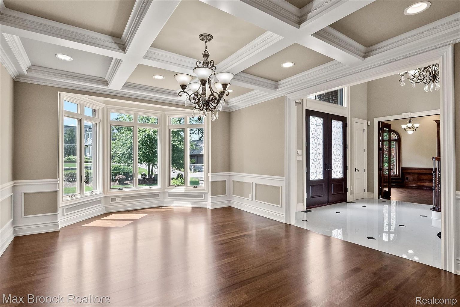 This is an interior shot of a dining room featuring a bay window, hardwood floors, and a coffered ceiling with a chandelier. The room is painted in neutral tones with white wainscoting, creating an elegant and inviting atmosphere. The view through the doorway shows a glimpse of the entryway with marble floors.