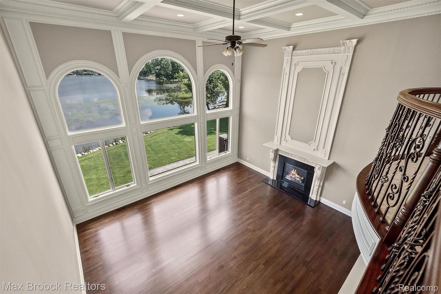 This is an interior shot of a luxurious living room featuring a wall of large arched windows overlooking a lake and lush greenery. The room boasts dark hardwood floors, a decorative fireplace with an ornate mantel, and a coffered ceiling with a ceiling fan. A curved staircase with wrought iron railings is visible on the right, adding to the room's grandeur.