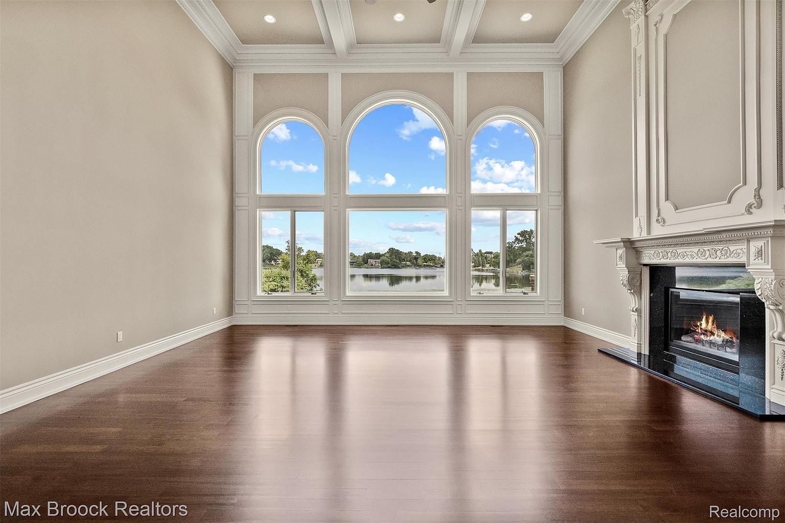 This is an interior shot of a luxurious living room featuring a grand fireplace and a wall of large arched windows offering a view of a lake. The room has hardwood floors, neutral-colored walls, and detailed white trim, creating an elegant and spacious atmosphere. The high ceilings and natural light enhance the room's appeal, making it a desirable space for relaxation and entertainment.