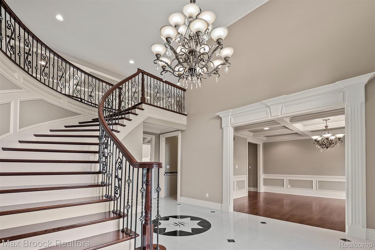 This grand foyer features a curved staircase with dark wood treads and ornate iron railings, leading to an upper level. A large, decorative chandelier hangs from the high ceiling, illuminating the space. The flooring is a mix of white and black tiles with a starburst pattern, and an open doorway leads to a dining room with coffered ceilings, creating an elegant and inviting entrance.