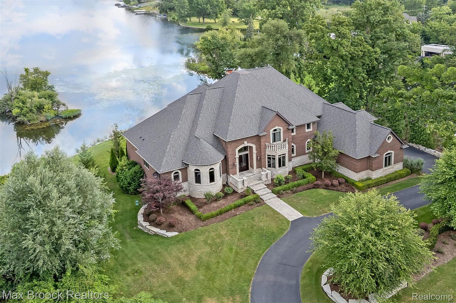 This aerial view showcases a luxurious brick home with a complex roofline and meticulously landscaped grounds. A winding driveway leads to the residence, which is situated on a waterfront property with a small island nearby. The overall impression is one of elegance and privacy, highlighting the property's prime location and upscale features.
