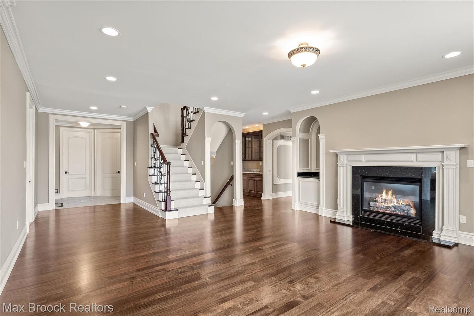 This is an interior shot of a living room featuring hardwood floors, a fireplace with a white mantle, and a staircase with wrought iron railings. The room is painted in neutral tones, and the lighting is bright, creating a warm and inviting atmosphere. The perspective is from the entrance of the room, showcasing the open layout and architectural details.