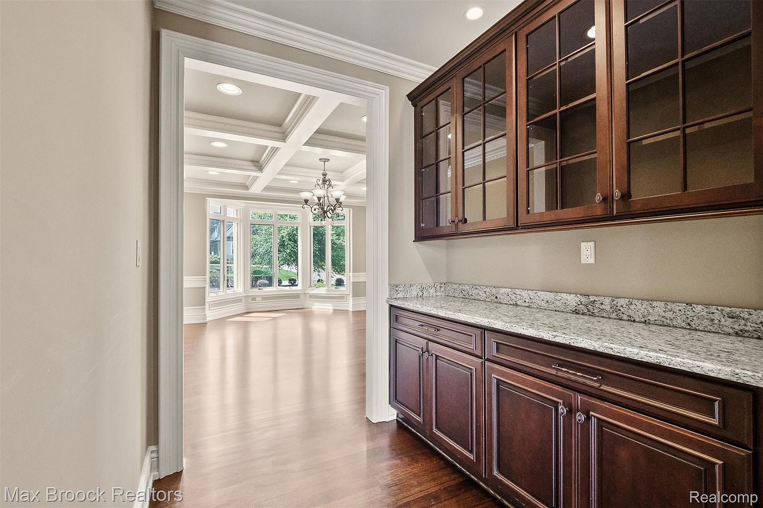 This interior shot showcases a dining room with dark wood cabinetry and a granite countertop, leading into a bright, open dining area with a coffered ceiling and a bay window. The transition between the spaces is seamless, highlighting the home's elegant design and ample natural light. The dark wood floors add warmth and sophistication to the space.
