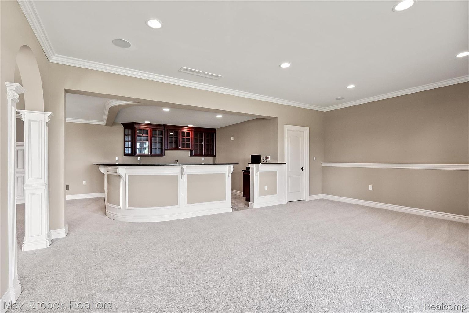 This is a spacious basement area featuring a wet bar with dark wood cabinets and a black countertop. The room is carpeted in a light gray, and the walls are painted in a neutral beige tone with white trim. Recessed lighting provides ample illumination, and the overall impression is clean and inviting.