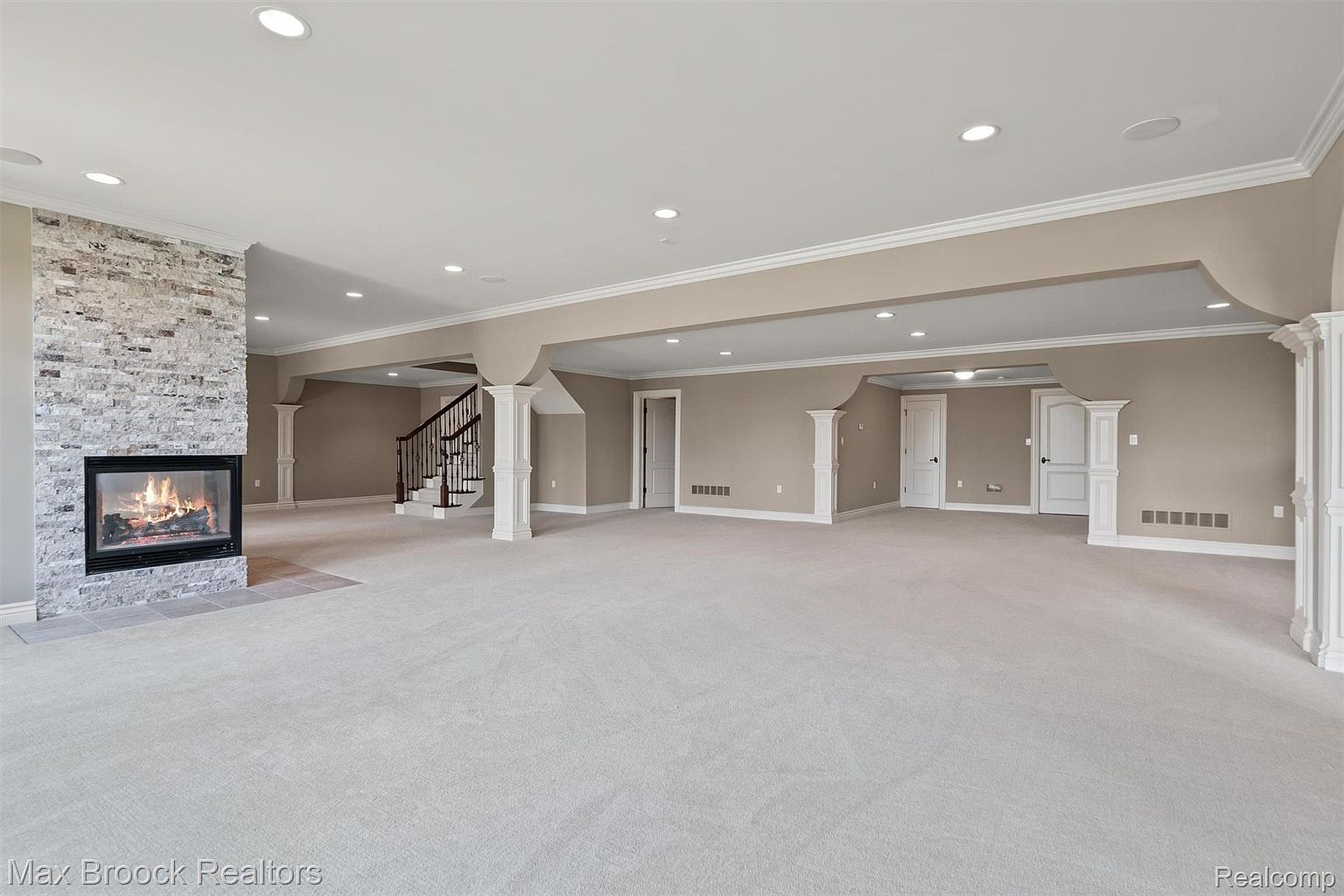 This is a spacious finished basement featuring a stone fireplace, neutral carpeting, and ample lighting from recessed fixtures. The area includes support columns and access to stairs, suggesting a versatile space for recreation or entertainment. The overall impression is clean, modern, and inviting.
