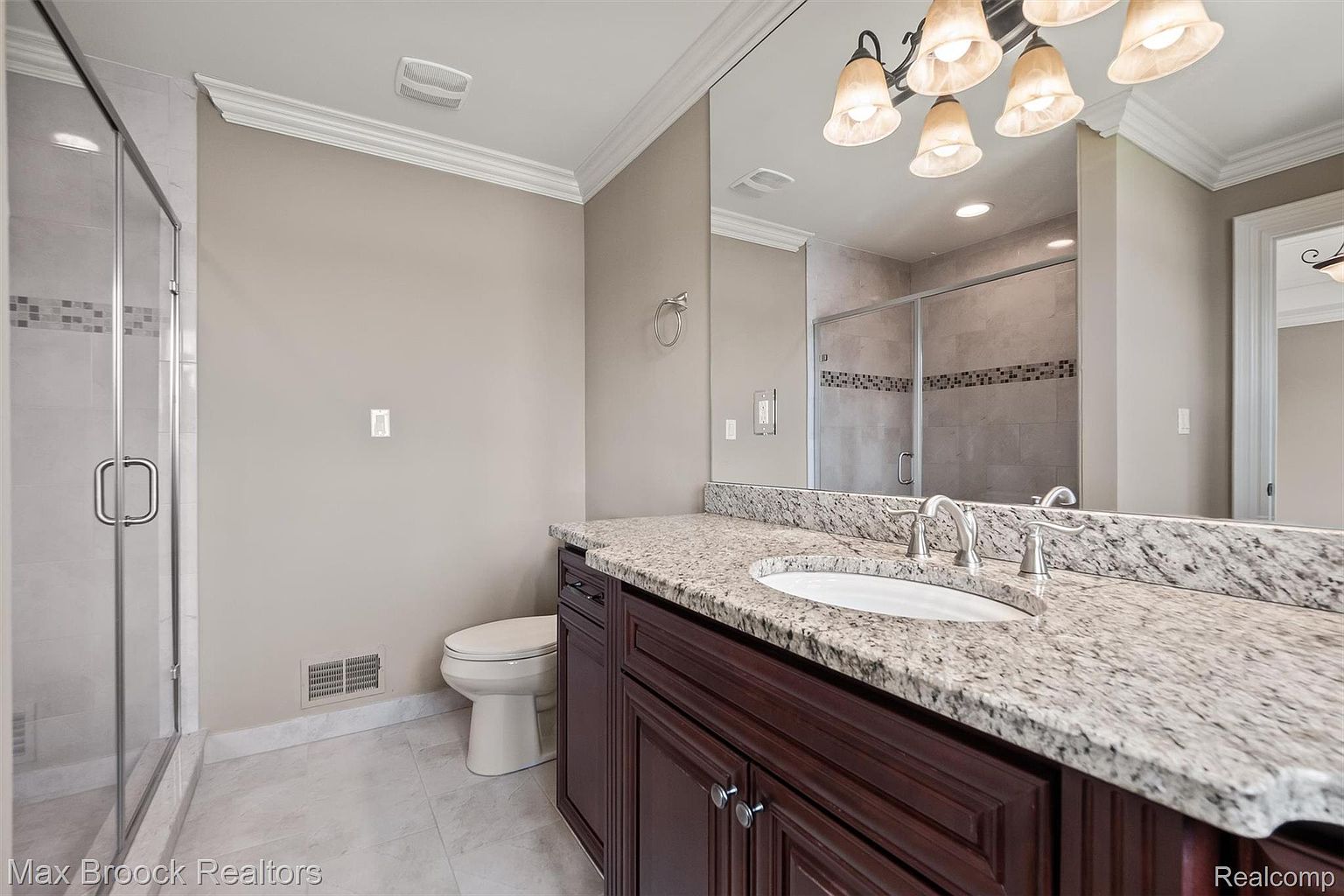This is a well-lit primary bathroom featuring a granite countertop vanity with dark wood cabinetry and a white sink. A large mirror reflects the space, including a glass-enclosed shower. The walls are painted in a neutral tone, and the flooring appears to be light-colored tile, creating a clean and inviting atmosphere.