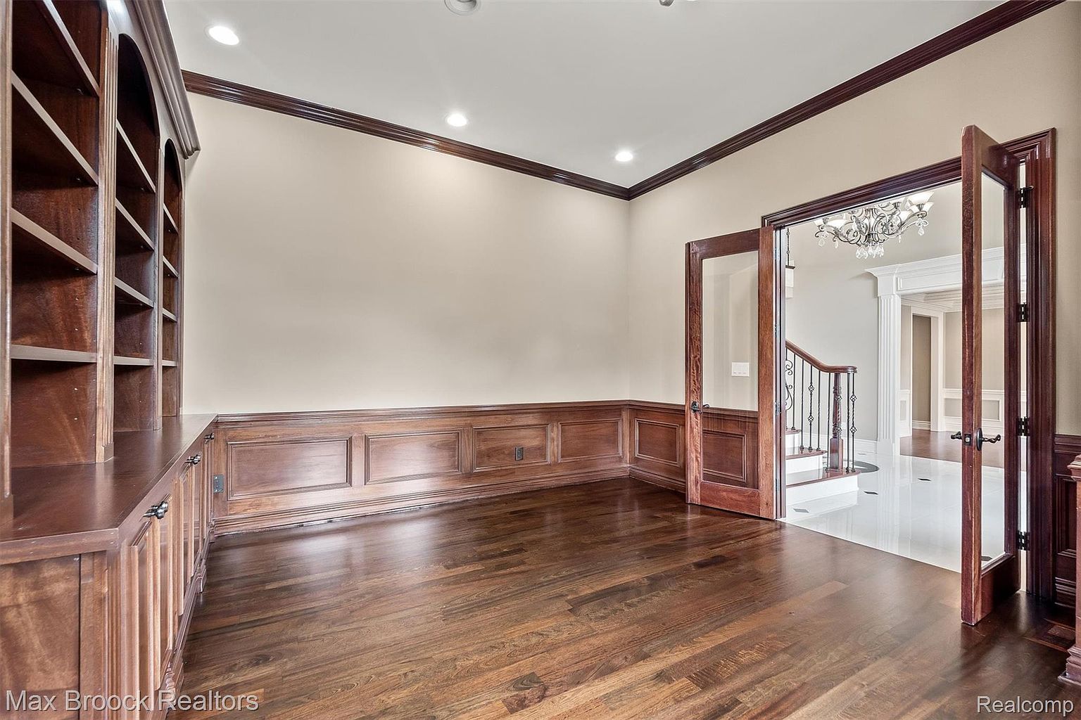 This is an interior shot of a home office or study, featuring rich wood paneling and built-in bookshelves. The room has hardwood floors and a neutral wall color, complemented by dark wood trim. Double glass doors lead to another room, suggesting a spacious and well-appointed home.