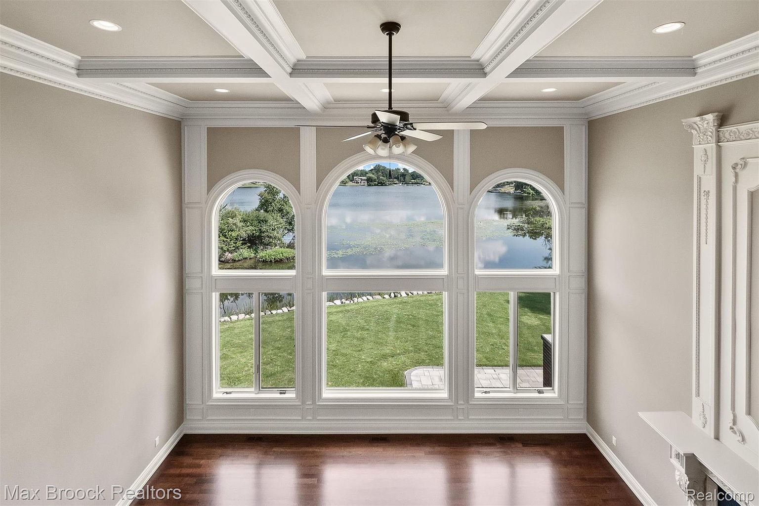 This sunroom features three large arched windows offering a view of a lake and lush greenery. The room is decorated with white trim and a coffered ceiling with a ceiling fan. The dark hardwood floors add warmth to the space, creating a bright and inviting atmosphere.