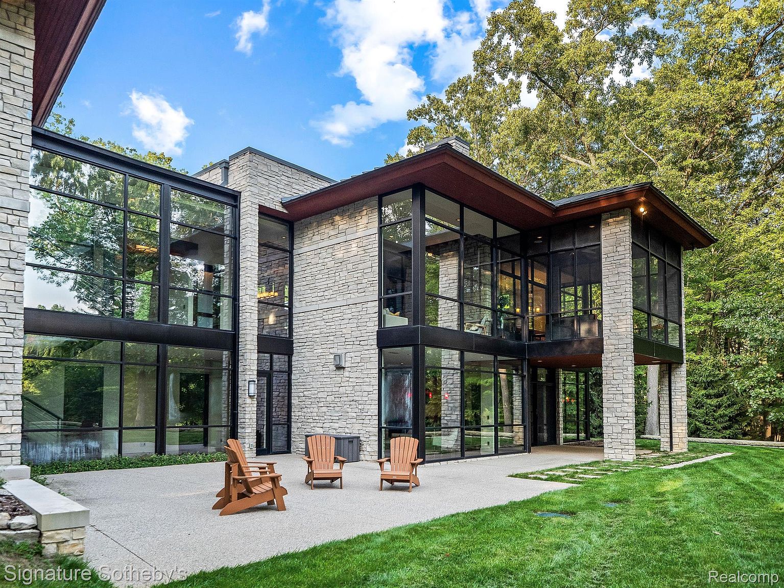 This is a rear exterior view of a modern, luxurious home. The architecture features a combination of stone and large glass windows, creating a bright and open feel. The outdoor space includes a patio area with wooden chairs and a well-maintained lawn, blending seamlessly with the surrounding trees.