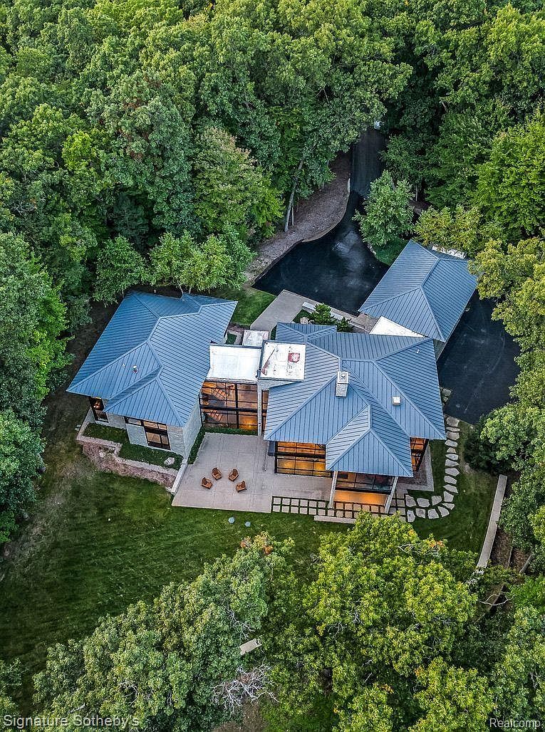 This aerial view showcases a modern home nestled within a lush, green forest. The house features a striking blue metal roof, large glass windows, and a spacious patio area with outdoor seating. A winding driveway leads to the property, adding to its secluded and private feel.