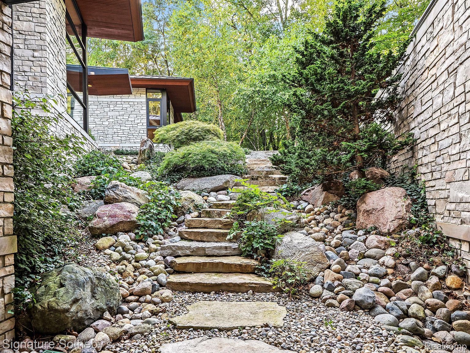This image showcases a beautifully landscaped yard with stone steps leading upwards through a rock garden. The steps are flanked by various sized rocks and pebbles, with lush greenery and trees providing a natural backdrop. The stone and plant life create a serene and inviting outdoor space.