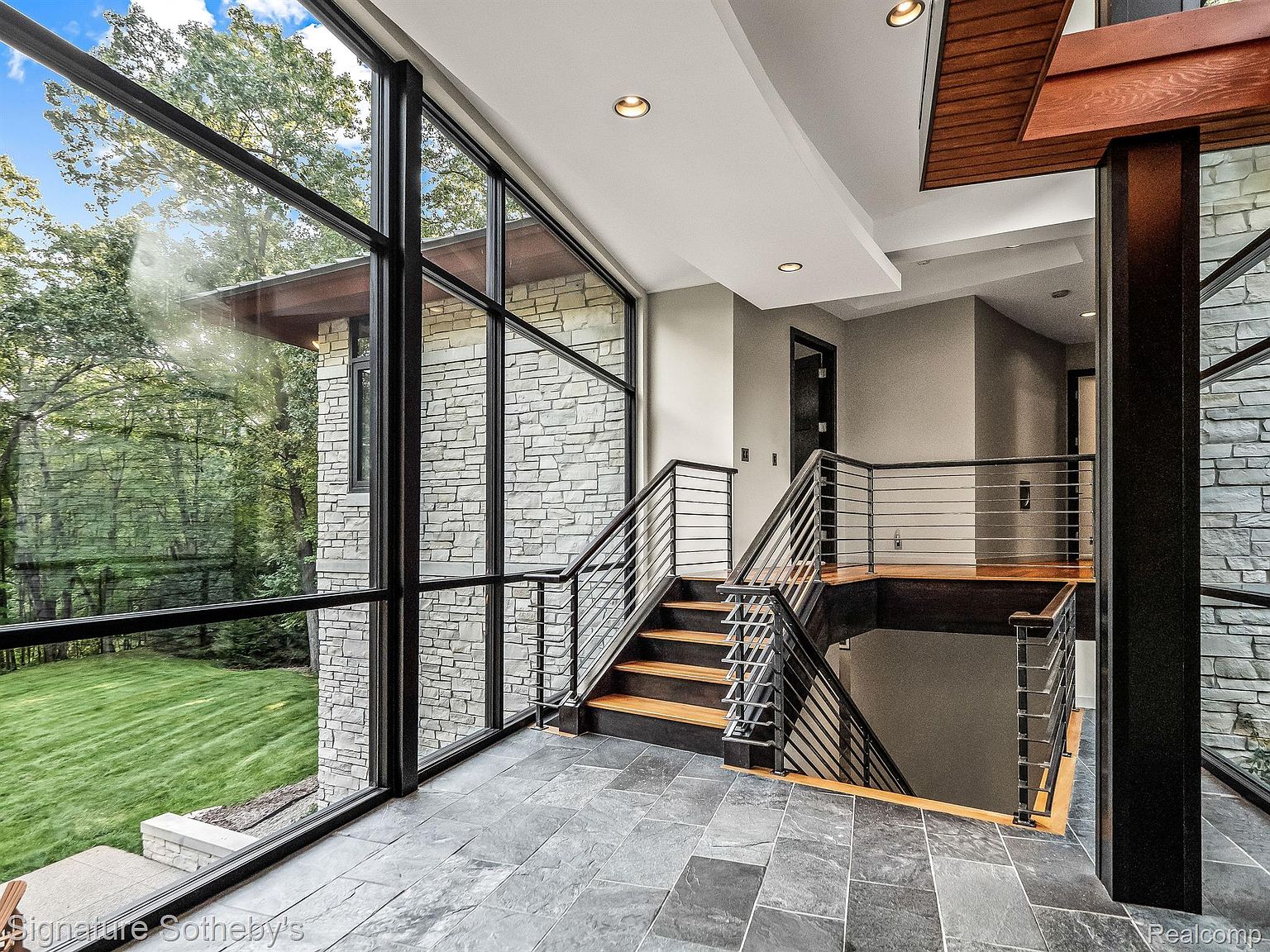 This interior shot showcases a modern hallway and staircase with a striking architectural design. The space features a combination of stone walls, large windows offering views of the exterior greenery, and a sleek metal railing system. The staircase leads to a lower level, and the overall aesthetic is contemporary and sophisticated, emphasizing natural light and clean lines.