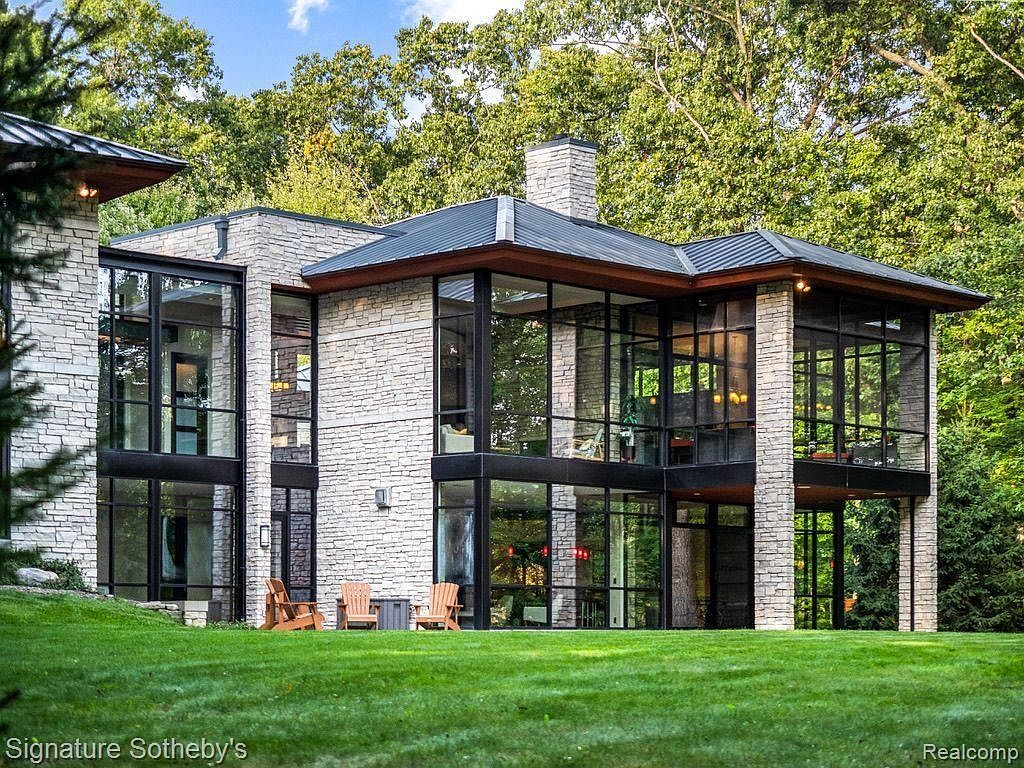 This is a striking front view of a modern, two-story home with a stone facade and expansive windows. The architectural design emphasizes clean lines and natural light, with a metal roof and well-manicured lawn adding to the property's curb appeal. The home is nestled among mature trees, providing a sense of privacy and tranquility.