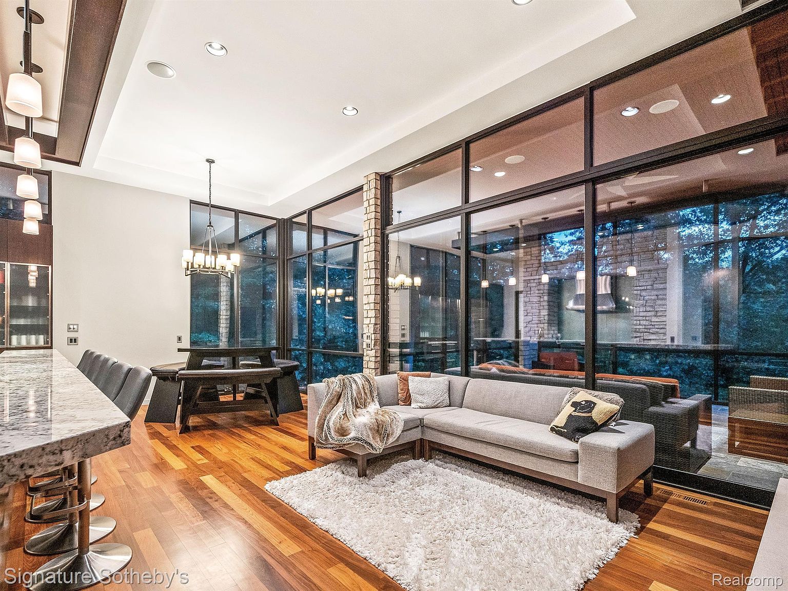 This is an interior shot of a modern living room featuring floor-to-ceiling windows that offer an expansive view of the outdoors. The room is furnished with a sectional sofa, a white shag rug, and a dining table with chairs. The hardwood floors add warmth to the space, while the large windows provide ample natural light, creating an inviting and luxurious atmosphere.