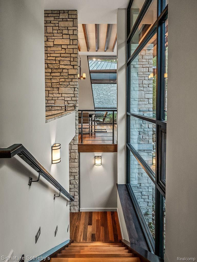 This interior shot showcases a modern staircase with wooden steps and a dark metal railing. A stone accent wall adds texture and visual interest, while sconce lighting provides a warm glow. Large windows offer natural light and a view to the outside, creating a bright and inviting space.