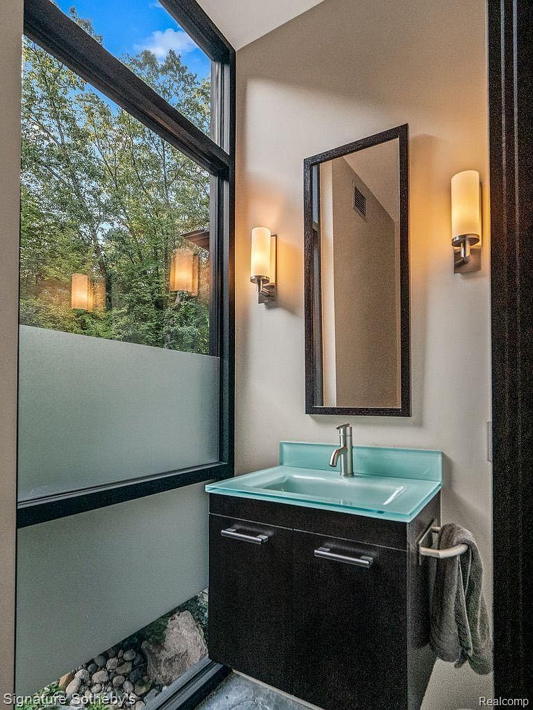 This is a modern bathroom featuring a unique design with a frosted window offering privacy and natural light. The vanity has a sleek, dark wood cabinet with a light blue glass countertop and sink. A rectangular mirror hangs above the vanity, flanked by sconce lighting, and there's a towel rack on the side. The floor has decorative rocks under the window.