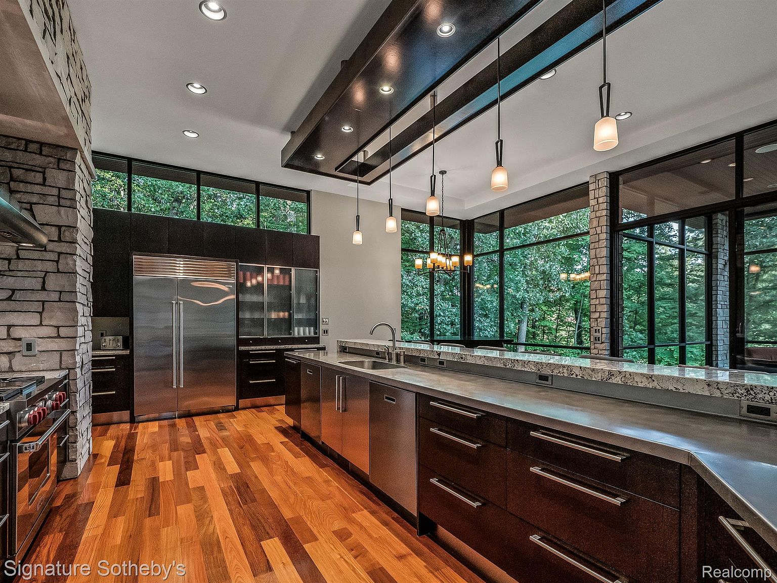 This is a modern kitchen with dark wood cabinetry, stainless steel appliances, and a long countertop with a stone backsplash. Large windows offer a view of the surrounding greenery, and pendant lights hang from a dark wood beam structure above the island. The kitchen features hardwood floors and a stone accent wall, creating a luxurious and contemporary feel.