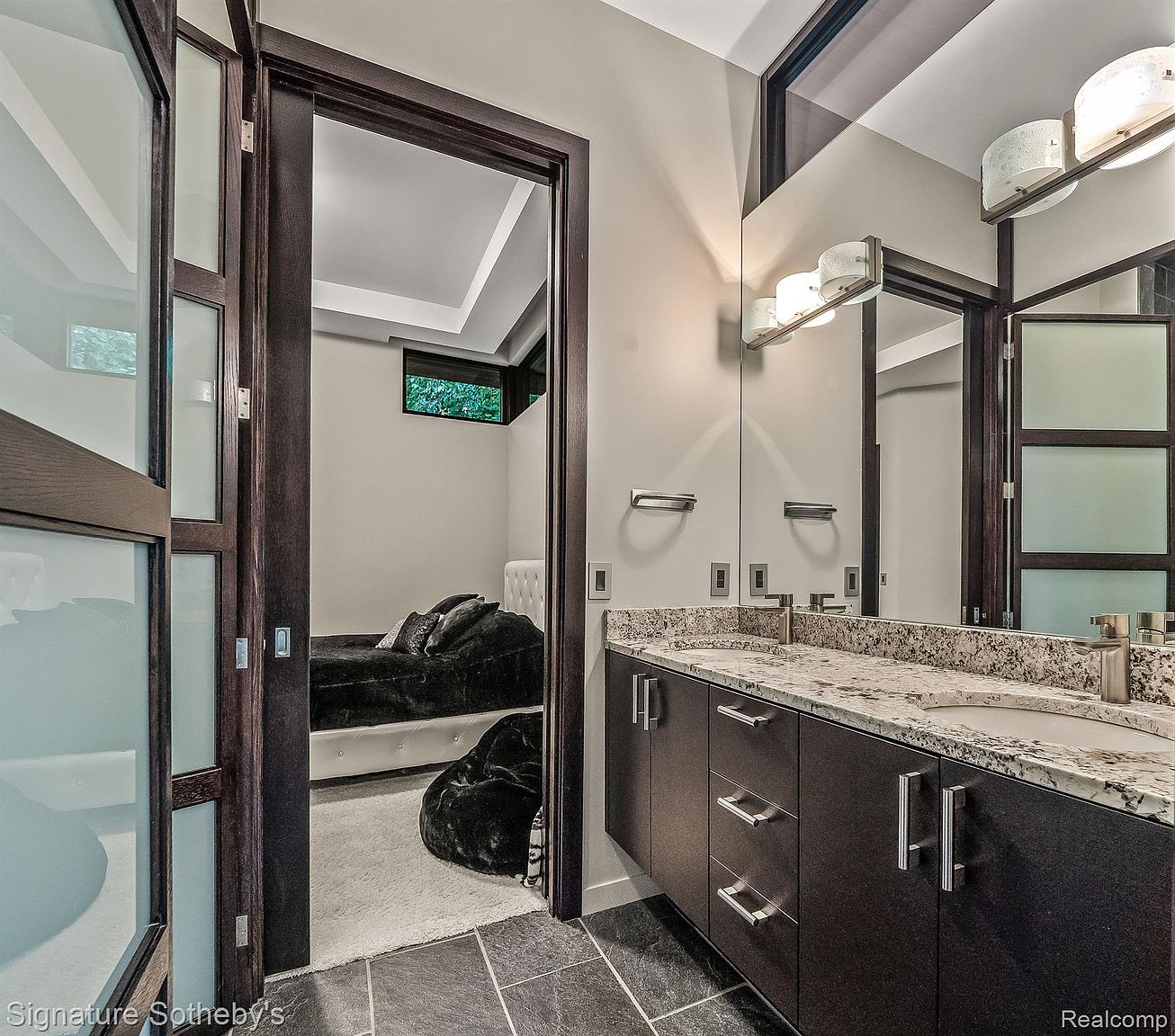 This image showcases a modern bathroom with a dark wood vanity topped with a granite countertop and dual sinks. A large mirror reflects the vanity and a glimpse into an adjacent bedroom, adding depth to the space. The bathroom features dark tile flooring and contemporary lighting fixtures, creating a sleek and stylish atmosphere.
