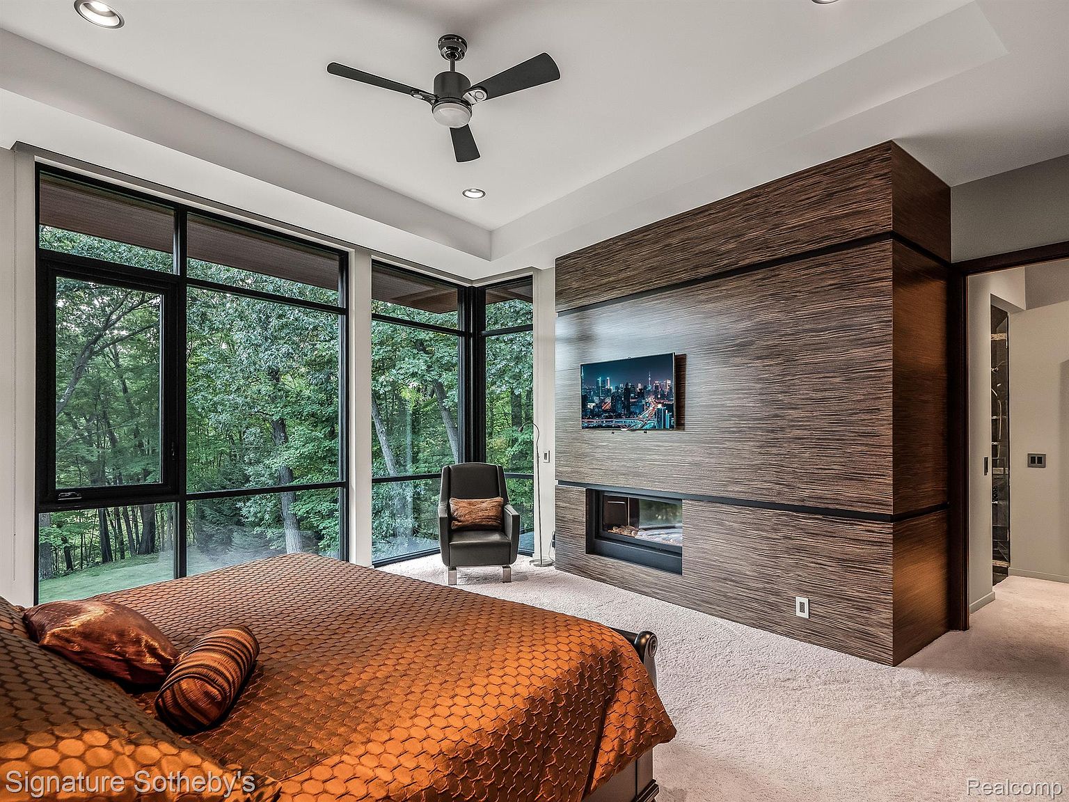 This is a luxurious primary bedroom featuring a large bed with an orange patterned comforter, floor-to-ceiling windows offering a view of a lush green forest, and a modern fireplace with a mounted television above it. The room is decorated in neutral tones with wood accents, creating a serene and sophisticated atmosphere, perfect for relaxation and comfort.