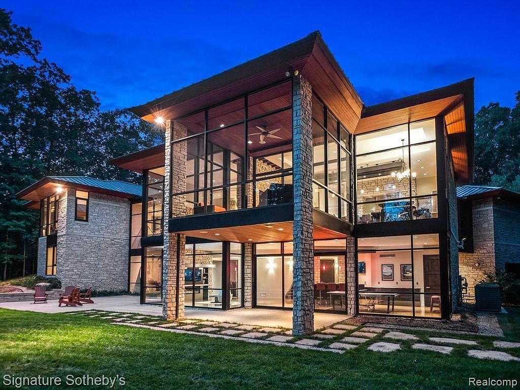This is a stunning front exterior view of a modern, multi-story home at dusk. The house features a unique architectural design with a combination of stone and large glass windows, creating a bright and open feel. The well-manicured lawn and stone pathway add to the property's curb appeal.