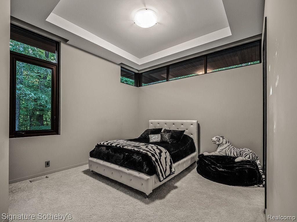 This is a bedroom featuring a white upholstered bed with black bedding and a zebra-striped throw. A decorative white tiger statue rests on a black beanbag near the bed. The room has a modern design with a unique ceiling feature and horizontal windows providing natural light.
