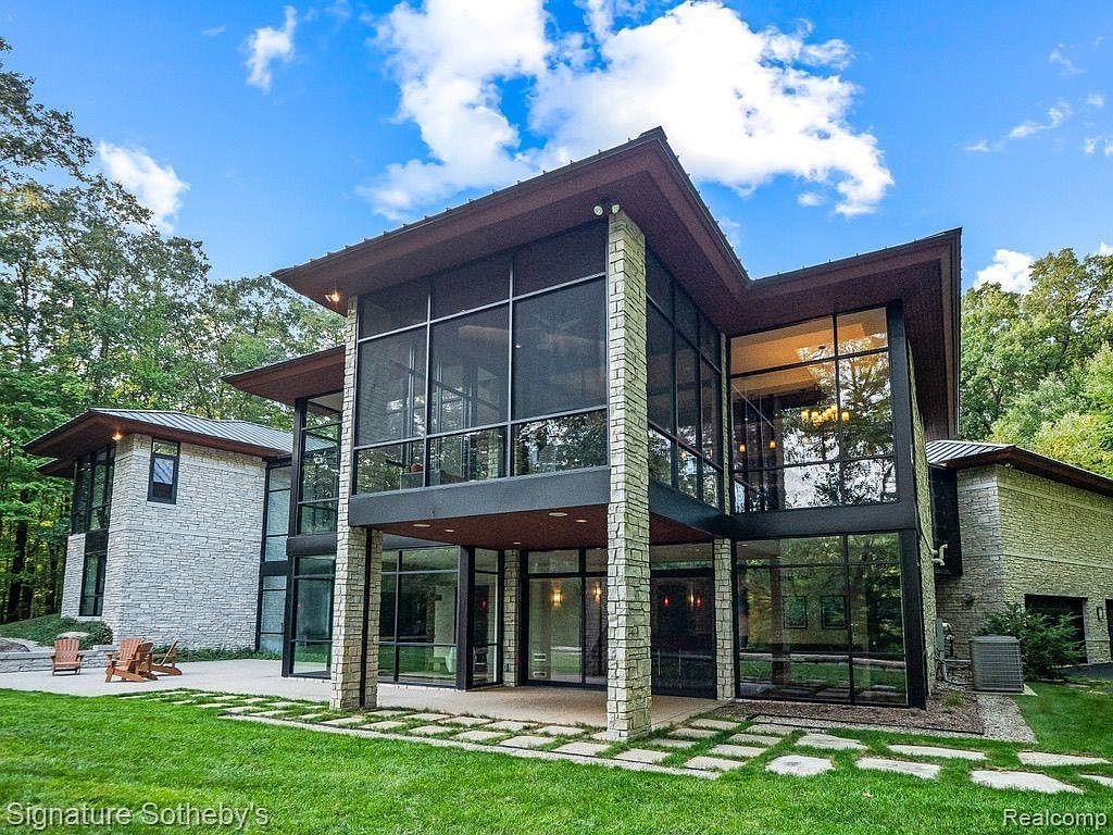 This is a front exterior view of a modern, luxurious home. The house features a combination of stone and glass, with large windows and a unique roofline. The landscaping is well-maintained with a green lawn and stone pathway, creating an inviting and sophisticated curb appeal.