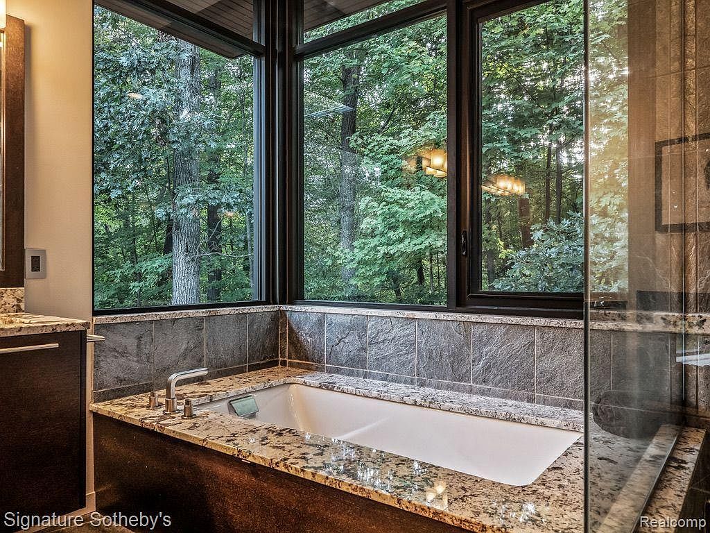 This is a luxurious primary bathroom featuring a large soaking tub positioned in front of a expansive window offering a view of a lush forest. The tub is surrounded by a granite countertop and dark wood cabinetry, complemented by gray stone tiling on the walls. A glass shower enclosure is visible to the right, enhancing the spa-like atmosphere.