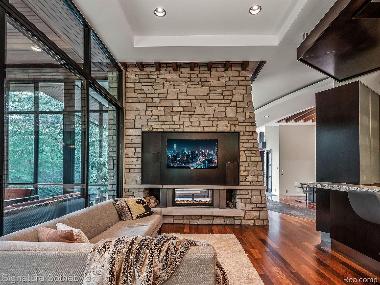 This is an interior shot of a modern living room featuring a stone fireplace with a built-in television. The room has large windows offering natural light and views of the surrounding greenery. A comfortable sectional sofa with throw pillows and a soft rug create a cozy and inviting atmosphere.