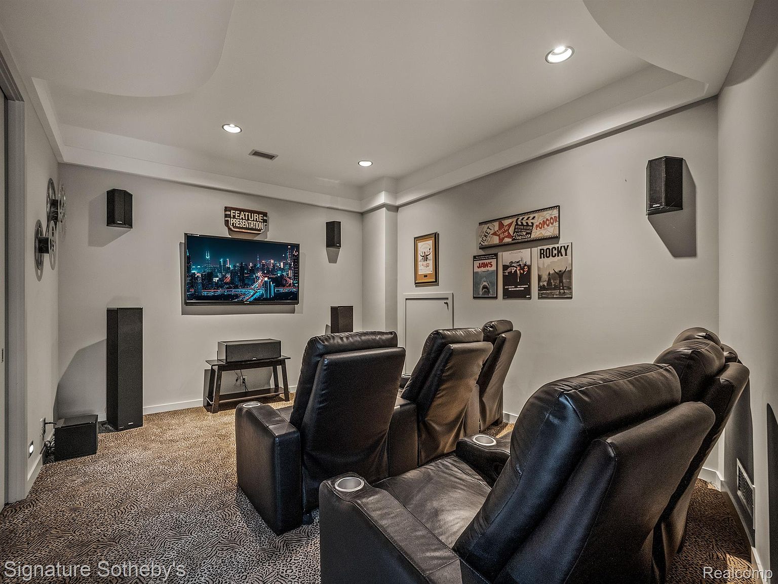 This is an interior shot of a home theater room. The room features multiple black leather recliner chairs, a large screen TV displaying a cityscape, and movie-themed decor on the walls. The room is well-lit with recessed lighting and has a cozy, inviting atmosphere, perfect for entertainment.