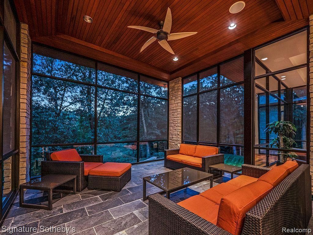 This is a sunroom featuring dark wicker furniture with orange cushions, offering a cozy and inviting atmosphere. Large windows provide ample natural light and views of the surrounding greenery. The ceiling is wood paneled with a ceiling fan, and the floor is tiled with large slate tiles, adding to the room's sophisticated yet relaxed ambiance.