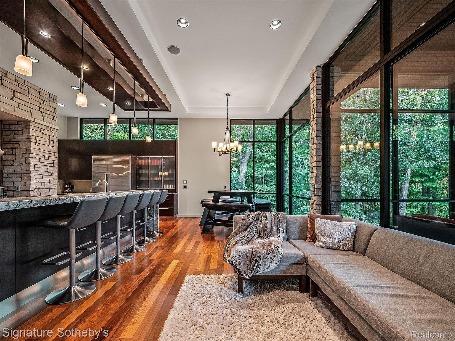 This interior shot showcases a modern living room and kitchen area with an open floor plan. The living room features a large sectional sofa, a plush rug, and floor-to-ceiling windows offering views of a lush forest. The kitchen area includes a stone-clad bar with seating and modern appliances, creating a seamless blend of living and dining spaces.