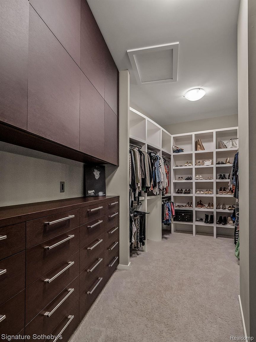 This is a well-organized walk-in closet featuring custom dark wood cabinetry with sleek, modern hardware. The closet includes ample hanging space and shelving, with a carpeted floor and recessed lighting. The overall impression is one of luxury and efficient storage.