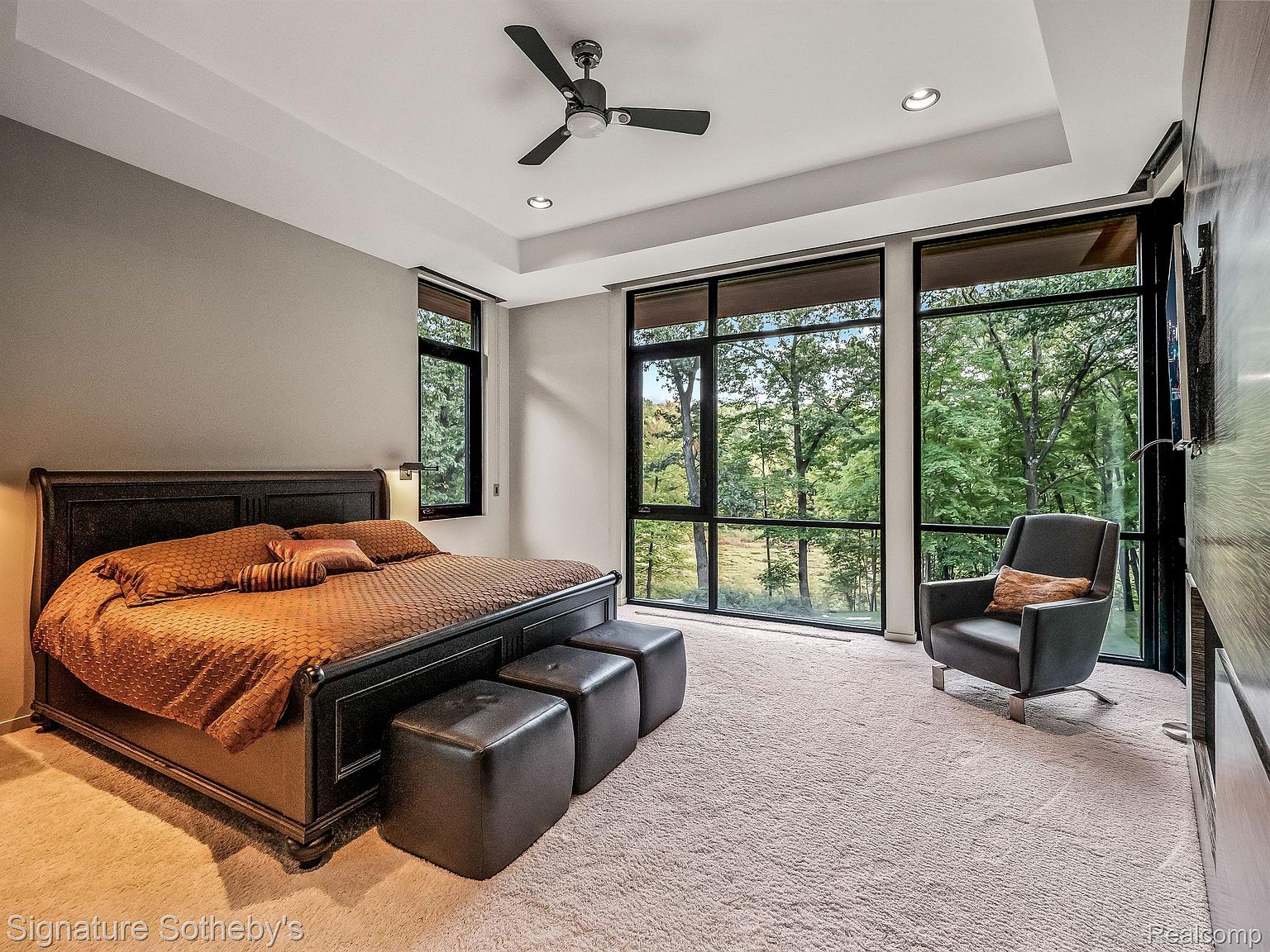 This is a luxurious primary bedroom featuring a dark wood bed frame with plush bedding and matching ottomans. Large windows offer a scenic view of the surrounding trees, enhancing the room's serene atmosphere. The room is carpeted and includes a modern armchair, creating a comfortable and stylish retreat.