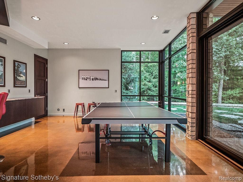 This interior shot showcases a spacious home gym featuring a ping pong table as the central element. Large windows offer views of the surrounding greenery, while the polished concrete floors reflect the natural light, creating a bright and inviting space. The room also includes built-in cabinetry and artwork, adding a touch of sophistication to the recreational area.