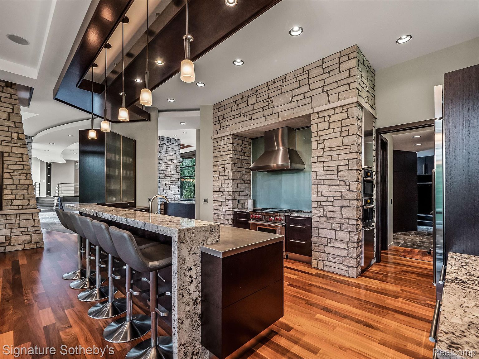 This is a modern, high-end kitchen featuring a large island with bar seating, stone accents on the walls and island, and dark wood cabinetry. Stainless steel appliances and pendant lighting add to the luxurious feel. The open layout and hardwood floors create a warm and inviting space.