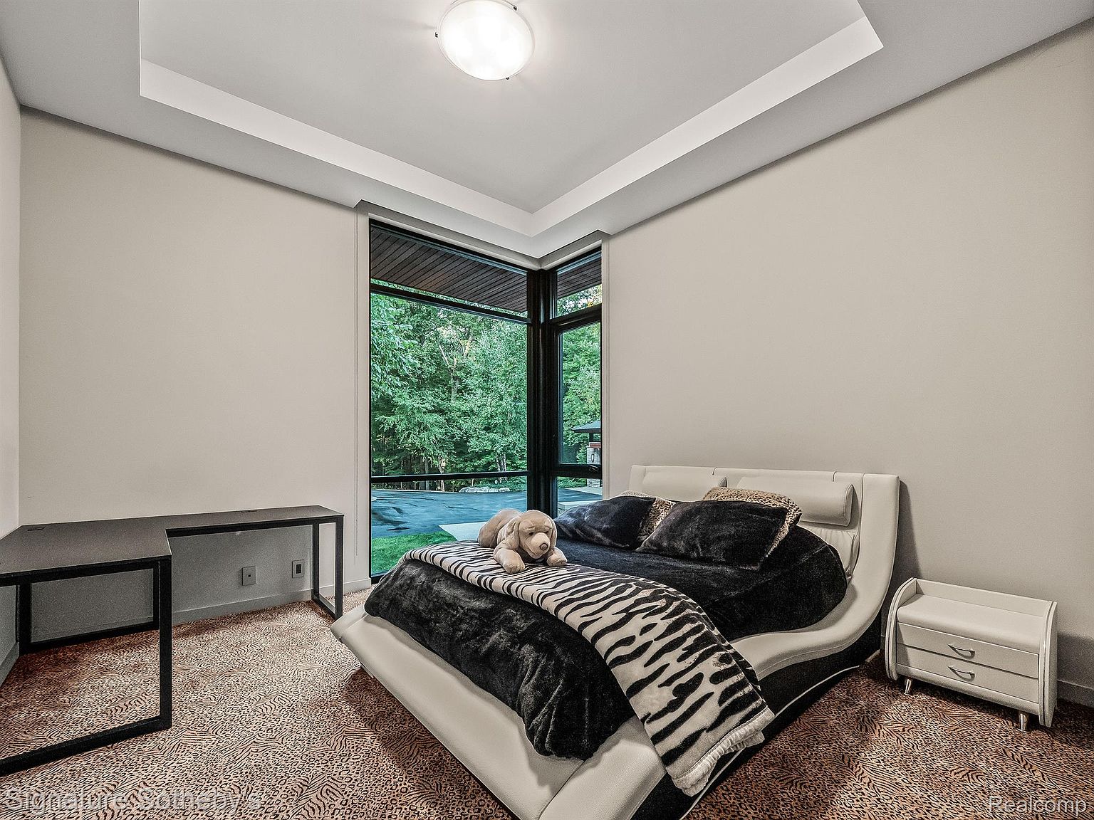 This bedroom features a modern aesthetic with a unique bed design, a zebra-striped throw, and a large window offering a view of the surrounding greenery. A sleek black desk sits against one wall, while a white nightstand complements the bed. The room is well-lit and appears spacious, creating a comfortable and stylish atmosphere.