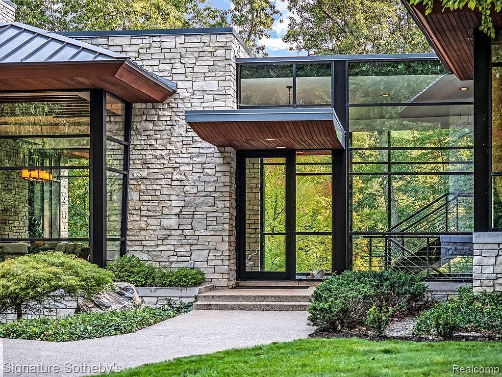 This image showcases the entryway of a modern home, emphasizing its architectural design. The entrance features a stone facade, large windows with black frames, and a wooden overhang, creating a striking visual appeal. The landscaping is well-maintained with greenery and a paved walkway leading to the front door, enhancing the property's curb appeal.
