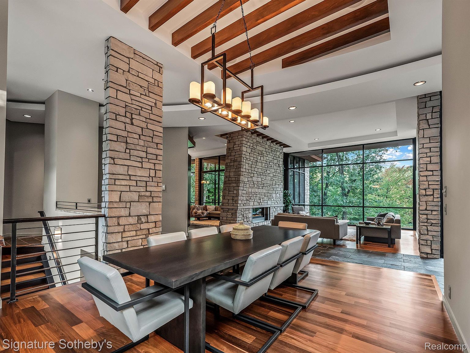 This interior shot showcases a sophisticated dining area with a large dark wood table surrounded by modern chairs with light upholstery. A striking linear chandelier hangs above the table, complementing the room's contemporary design. The space flows into a living area with a stone fireplace and large windows offering views of the outdoors, creating a seamless and inviting atmosphere.