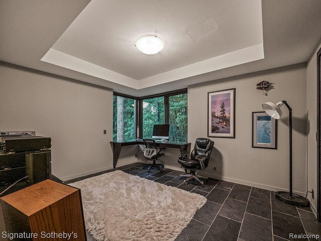 This is an interior shot of a home office featuring a corner desk setup with a computer, two office chairs, and natural light streaming through the windows. The room is decorated with framed artwork and a modern floor lamp, complemented by a plush rug on a dark tile floor. The overall aesthetic is contemporary and functional, suitable for a professional workspace.